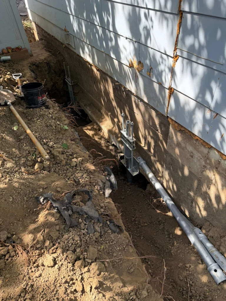 Multiple helical pier brackets in excavated access points along a foundation wall during stabilization work in Longmont by Fort August Foundation